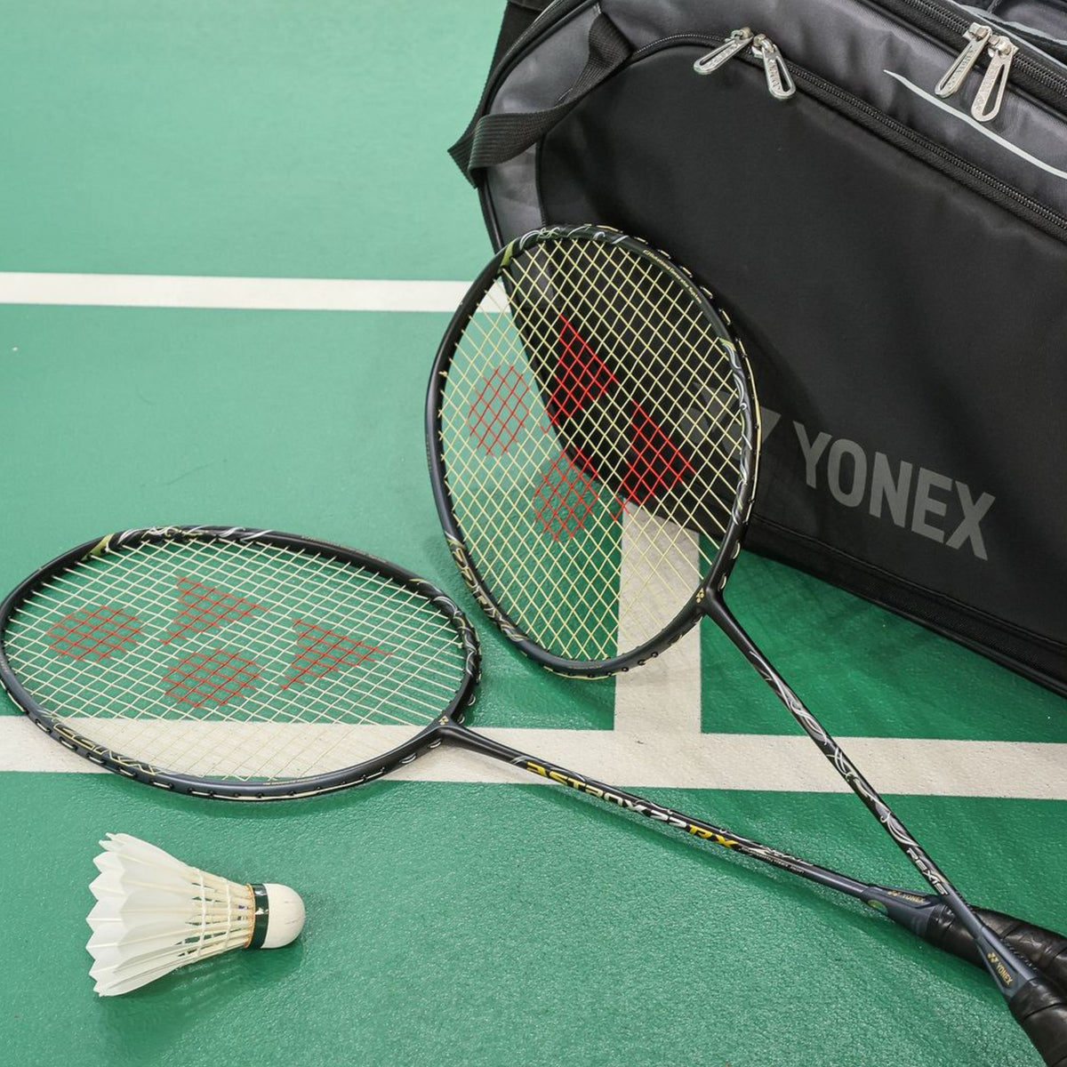 Two badminton rackets and a shuttlecock on a green court with a Yonex bag in the background.